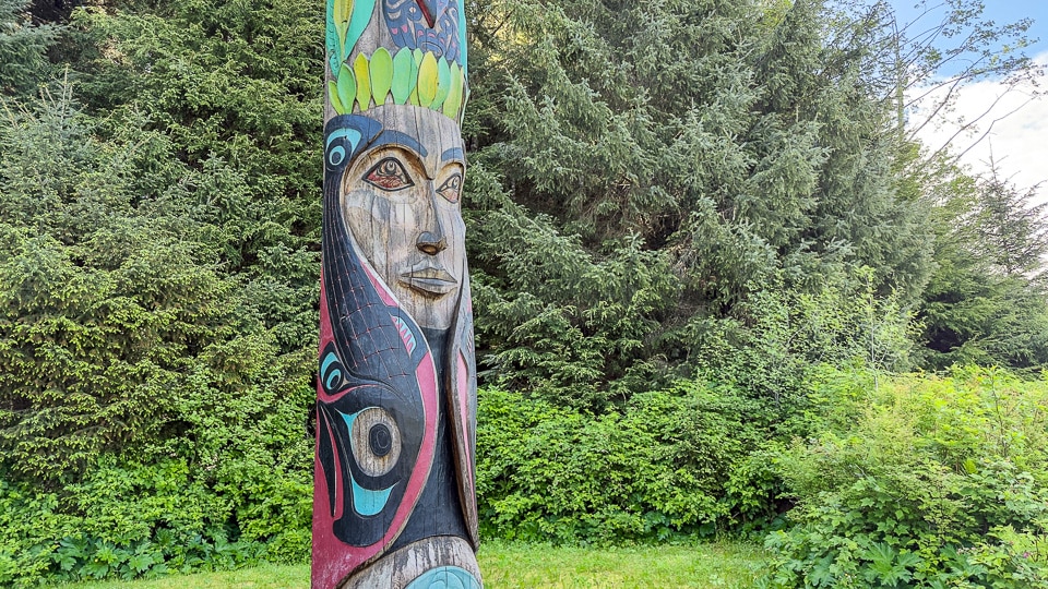 Close-up of a richly painted totem section at Sitka National Historical Park showing a human face with flowing black formline design, pink and turquoise accents, and green feather or wing elements at the top, with Sitka spruce forest behind