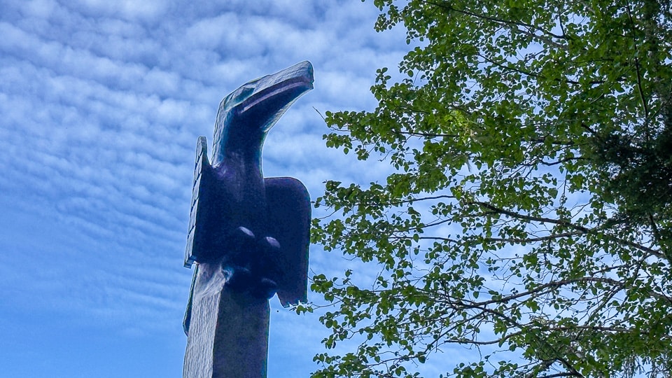 A dramatic low-angle view looking up at the top of a totem pole at Sitka National Historical Park, showing a large dark bird figure with an open beak silhouetted against a bright blue sky with scattered clouds and leafy tree branches