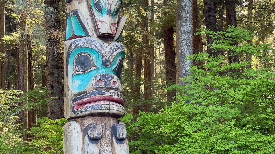 Close-up of two stacked figures on a totem pole at Sitka National Historical Park — a bold teal animal face with open mouth at top and a larger weathered teal face with prominent features below, set against green rainforest