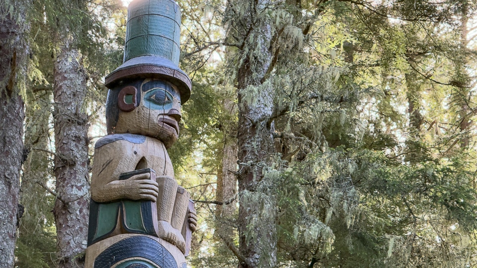 Close-up of the Yaadaas Crest Pole at Sitka National Historical Park showing a human figure wearing a tall green top hat and holding an object, surrounded by moss-draped spruce trees in dappled light