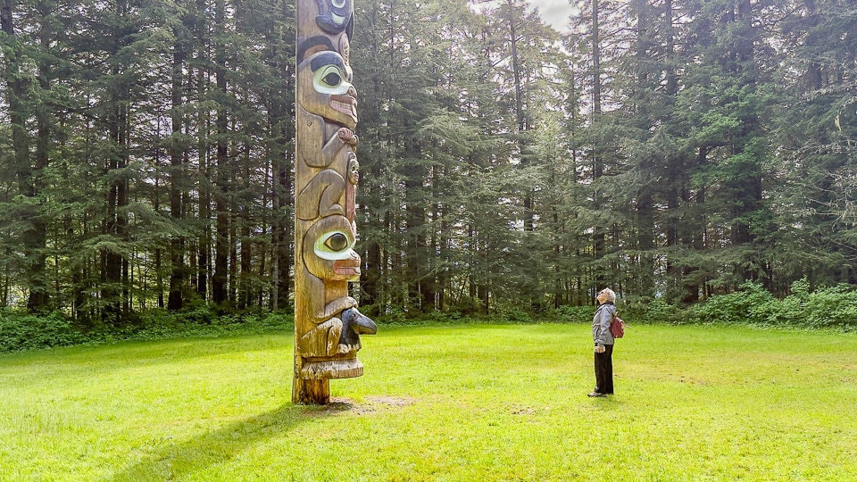 A woman looks up at the towering K'alyaan Pole in the grassy clearing at Sitka National Historical Park, surrounded by tall spruce trees — the pole's scale dwarfs the visitor standing beside it