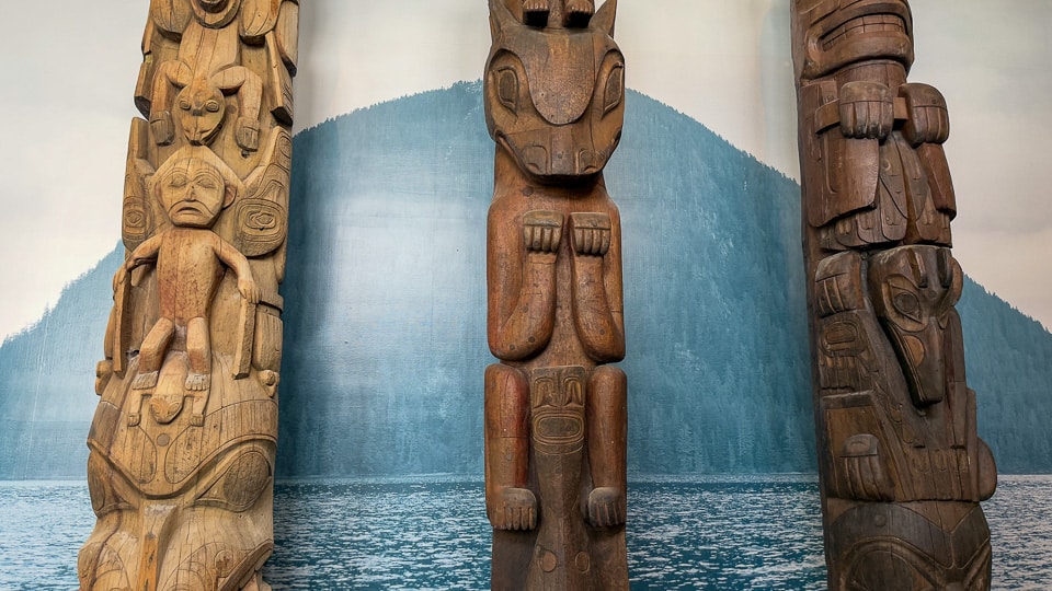 Three totem poles displayed inside Totem Hall at the Sitka National Historical Park visitor center, photographed against a painted mural of an Alaskan coastal mountain and water scene — the left pole shows a human figure with outstretched arms, the center pole features a large animal face, and the right pole displays stacked figures in darker wood