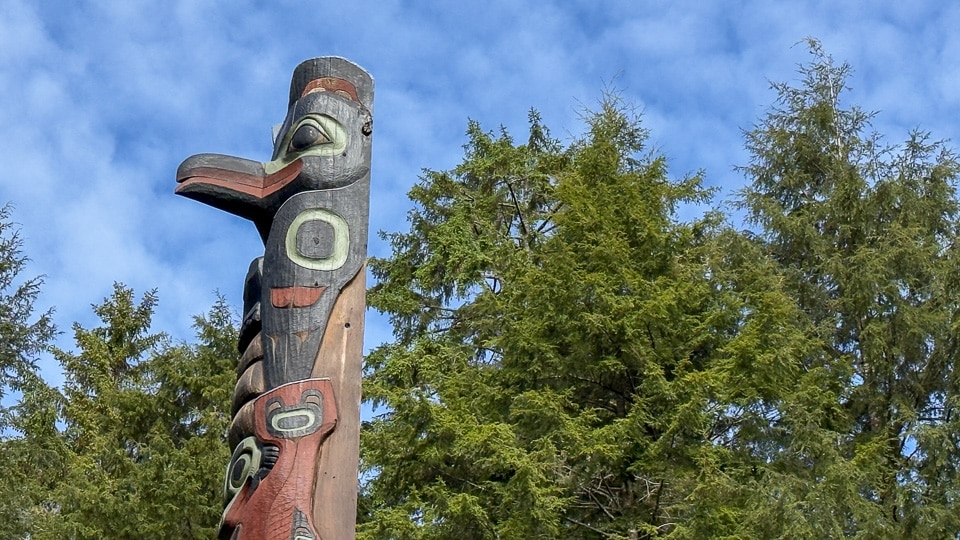 The upper section of a totem pole at Sitka National Historical Park showing a large bird head with an open beak and a bold circular eye at the top, and another figure below, photographed against a bright blue sky and spruce canopy