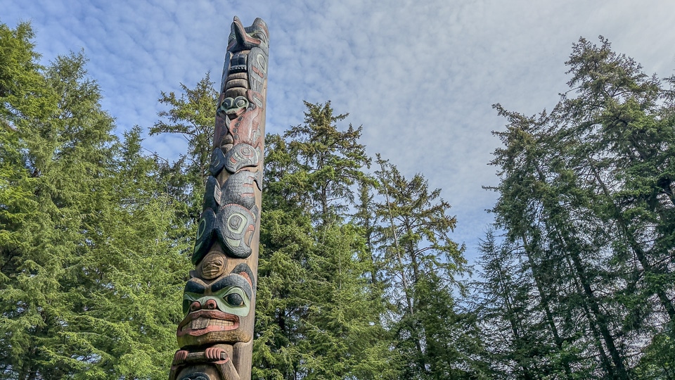 A tall totem pole at Sitka National Historical Park with multiple stacked faces and figures photographed from below against a dramatic sky with scattered clouds, with tall spruce trees rising on both sides