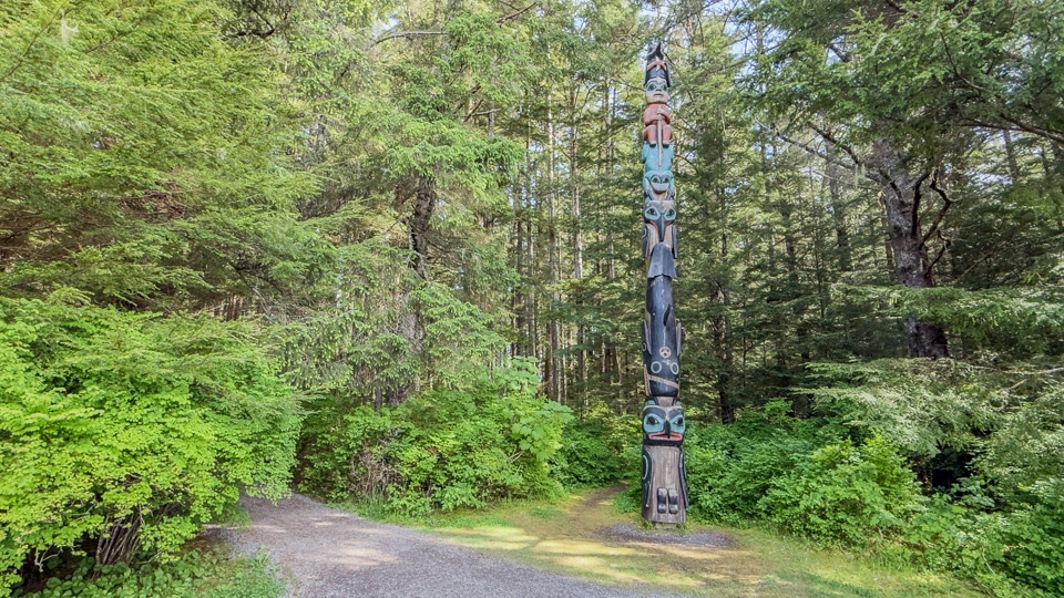 A wide view of the flat gravel Totem Trail at Sitka National Historical Park with a tall totem pole standing in a sunlit clearing surrounded by lush green rainforest.