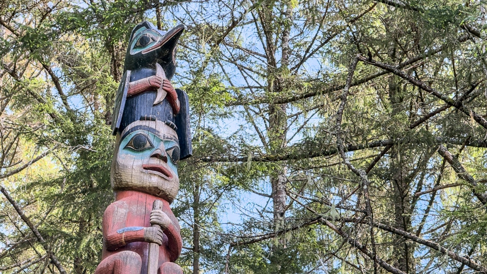 The upper section of a totem pole at Sitka National Historical Park showing a large raven at the top with a long open downward-curving beak, a human face below it, and a red figure beneath, set against tree branches and open sky