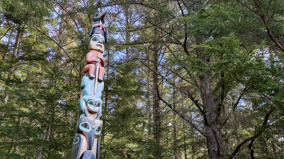 A full-length view of a tall multi-figure totem pole at Sitka National Historical Park rising above the forest canopy, with a bird figure at the top, a human face in red below, a large teal-eyed figure in the middle, and stacked figures at the base