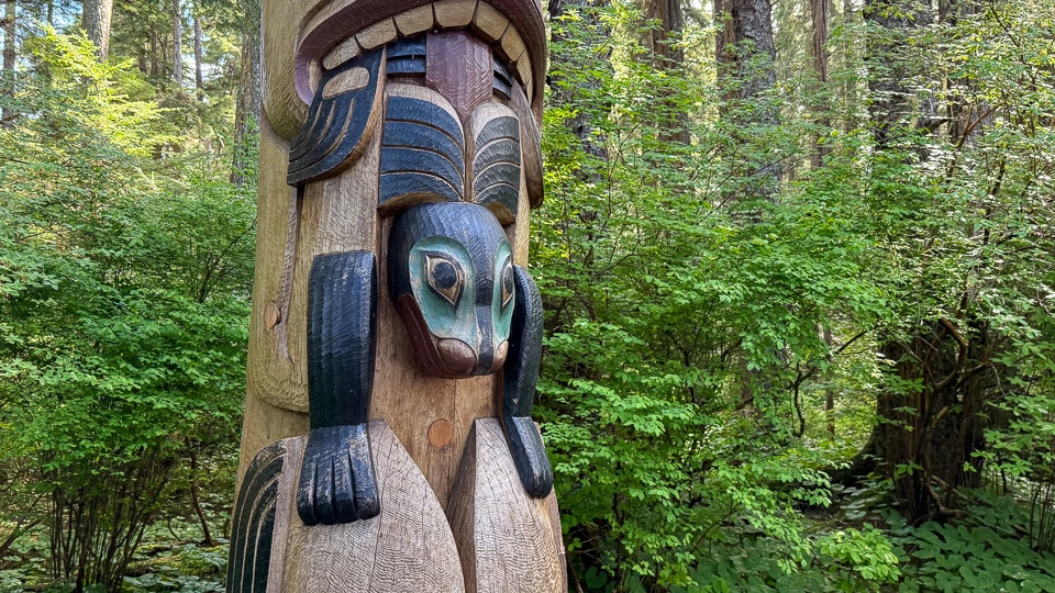 Close-up detail of a bird figure on a totem pole at Sitka National Historical Park, with distinctive teal round eyes and dark wings folded at its sides, with lush green forest undergrowth behind