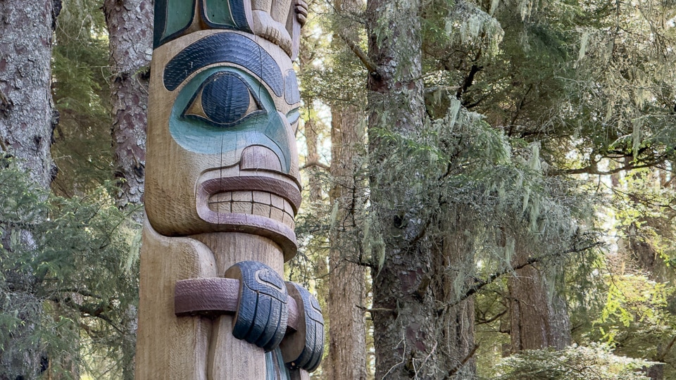 Close-up of a totem face carved in pale fresh cedar at Sitka National Historical Park, with large teal-green eyes, bared teeth, and a smaller dark figure below gripping the pole with claws, with rainforest trees behind