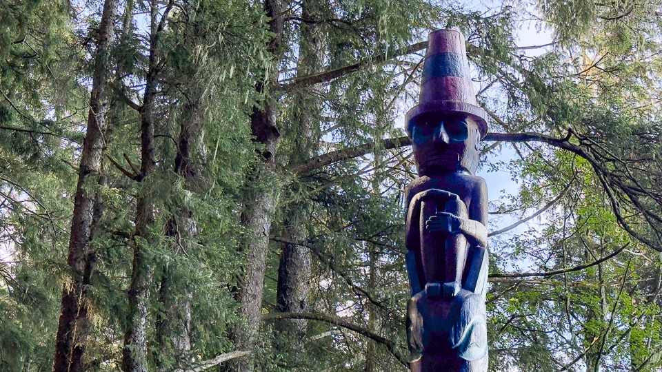 A slim dark totem pole at Sitka National Historical Park rising high above the rainforest canopy, topped by a carved human figure wearing a top hat, photographed from below looking up through the tree branches