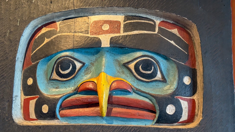 A boldly painted carved panel inside the Sitka National Historical Park visitor center showing a bird face with teal and blue coloring, a yellow beak, and red and white geometric formline design on a dark background
