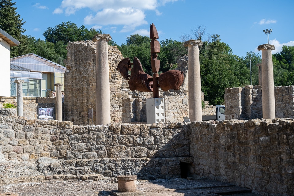 Stone walls and standing columns of the Villa Romana del Casale, with a modern rust-colored sculpture and the visitor building behind